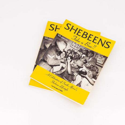 Book titled 'Shebeens Take a Bow' with yellow cover and black and white photo inside on a white background